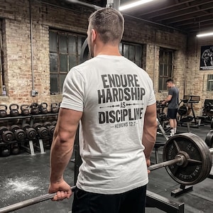 May include: A white short-sleeved t-shirt featuring black text on the back that reads: ENDURE HARDSHIP AS DISCIPLINE - HEBREWS 12:7. The shirt is worn by a person in a gym setting with weightlifting equipment visible in the background.