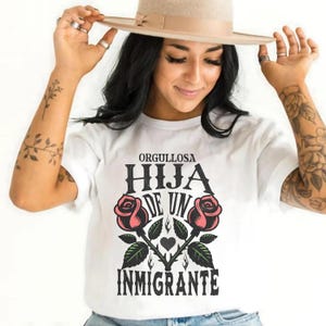 May include: White t-shirt with the text "ORGULLOSA HIJA DE UN INMIGRANTE" in black, with two pink roses and green leaves. The person in the image is wearing a beige hat and has tattoos on their arms.