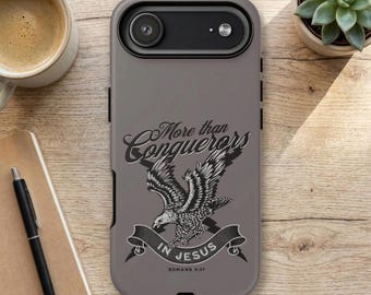 May include: A matte grey smartphone case featuring a black and white graphic of an eagle in flight. The text reads More than Conquerors IN JESUS and Romans 8:37. The case has a black camera housing at the top with two circular lens cutouts.