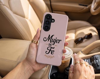 May include: A light pink smartphone case featuring the black text "Mujer DE Fe" in a stylized script font, accompanied by a small gold-colored decorative floral graphic at the bottom, with a triple-lens camera cutout visible at the top.