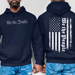 K&ouml;nnte beinhalten: Zwei marineblaue Kapuzenpullover mit einem wei&szlig;en, abgenutzten amerikanischen Flaggenmuster auf der R&uuml;ckseite. Ein Pullover hat den Text "We the People" in Wei&szlig; auf der Vorderseite.