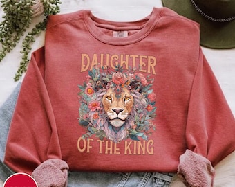 May include: A red sweatshirt with the text "Daughter of the King" and a floral design featuring a lion's head.