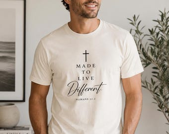 May include: A cream-colored short-sleeve t-shirt featuring a black graphic print. The design includes a cross symbol above the text "MADE TO LIVE Different" and the reference "ROMANS 12.2" printed in a combination of serif and script fonts.