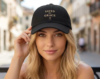 May include: A black baseball cap featuring the text SAVED BY GRACE embroidered in a light tan color on the front panel, positioned above a small cross symbol. The cap has a structured crown and a curved brim, with a classic six-panel design.