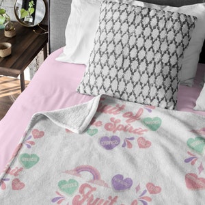May include: White fleece throw blanket with a rainbow and the words "Fruit of the Spirit" in pink and purple. The blanket is covered in pink, purple, and green hearts with words like "love", "joy", "peace", "patience", "kindness", "goodness", "faithfulness", "gentleness", and "self-control" printed on them.