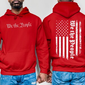 K&ouml;nnte beinhalten: Zwei rote Kapuzenpullover mit dem Aufdruck "We the People". Ein Pullover hat ein wei&szlig;es, abgenutztes amerikanisches Flaggenmuster auf der R&uuml;ckseite.