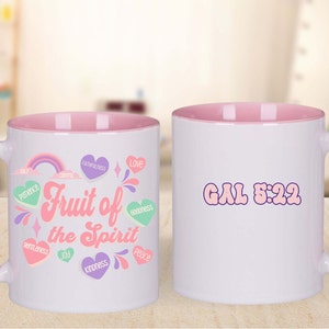 May include: White ceramic mug with a pink rim and handle. The mug features a colorful design with the words "Fruit of the Spirit" and a list of the fruits of the Spirit: love, joy, peace, patience, kindness, goodness, faithfulness, gentleness, and self-control. The back of the mug has the text "GAL 5:22" in pink.