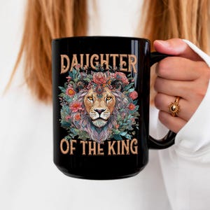 May include: Black coffee mug with a floral design and a lion's head in the center. The text "Daughter of the King" is printed on the mug.
