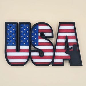 May include: Wooden USA sign with a patriotic theme. The letters are black with the American flag design incorporated. The letters "U" and "S" feature the blue field with white stars, while the "A" has red and white stripes and a silhouette of a saluting soldier.