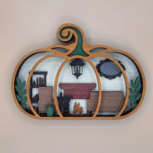 Peut inclure: Une décoration murale en bois superposé en forme de citrouille représentant une scène d'intérieur détaillée. Le design comprend une cheminée avec une flamme découpée, une lanterne suspendue, une étagère, un canapé, un fauteuil et des plantes en pot.