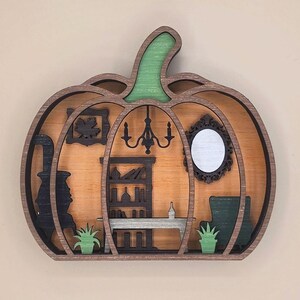 May include: A layered wooden pumpkin-shaped wall decoration featuring a miniature interior scene. The design includes a black stove, a bookshelf, a table, a chandelier, a mirror, a framed leaf, and two green potted plants against a light wood background.