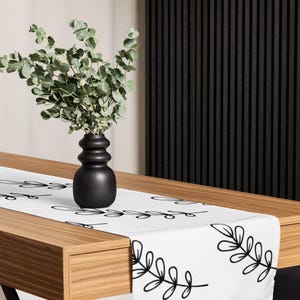 May include: A white table runner with a black leaf design. A black vase with a green plant sits on a wooden table. The table is in front of a black slatted wall.