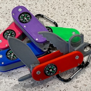 May include: Five multi-colored pocket knives with compass features. The knives are arranged in a pile with the blades open. The knives are red, blue, purple, green, and gray.