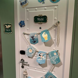 Puede incluir: Una puerta blanca con marco verde decorada con pegatinas de tem&aacute;tica n&aacute;utica y una cuerda de banderas. La puerta tiene una placa de n&uacute;mero con el n&uacute;mero 9614. Las banderas tienen im&aacute;genes de Mickey Mouse, un ancla de barco y helado.