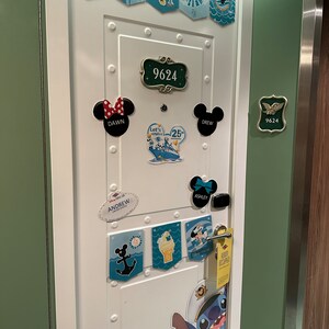 Puede incluir: Una puerta blanca con marco verde decorada con pegatinas y imanes con tem&aacute;tica de Disney. La puerta tiene una placa de n&uacute;mero que dice "9624". Hay varias pegatinas con nombres como "Dawn", "Drew", "Ashley" y "Andrew". Tambi&eacute;n hay pegatinas con im&aacute;genes de Mickey Mouse, Stitch y un crucero.