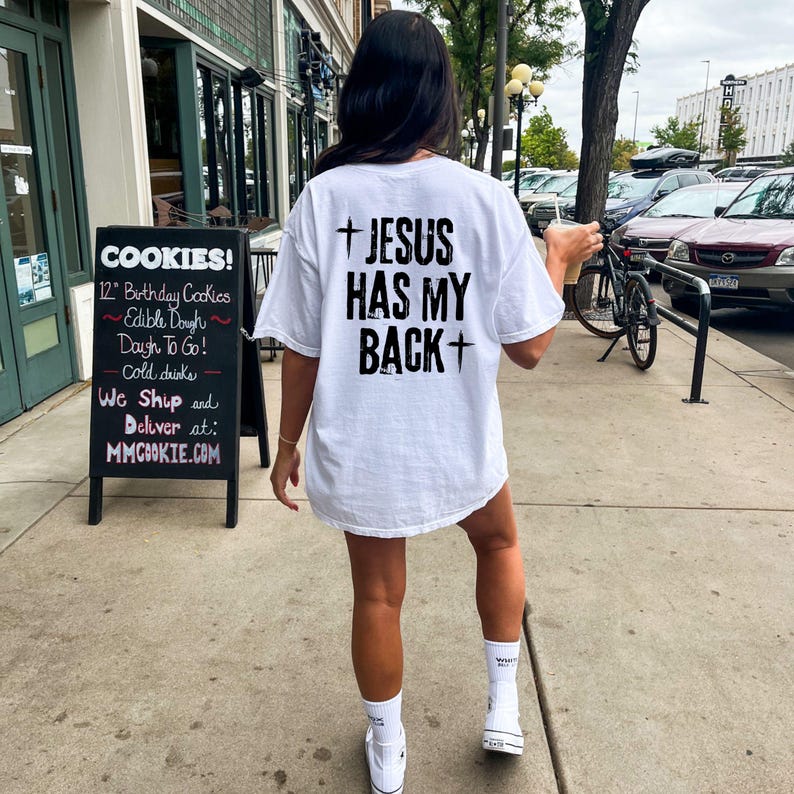 May include: White t-shirt with the text "JESUS HAS MY BACK" in black, with a cross above and below the text. The person is wearing white sneakers and socks. A sign for cookies is visible in the background.