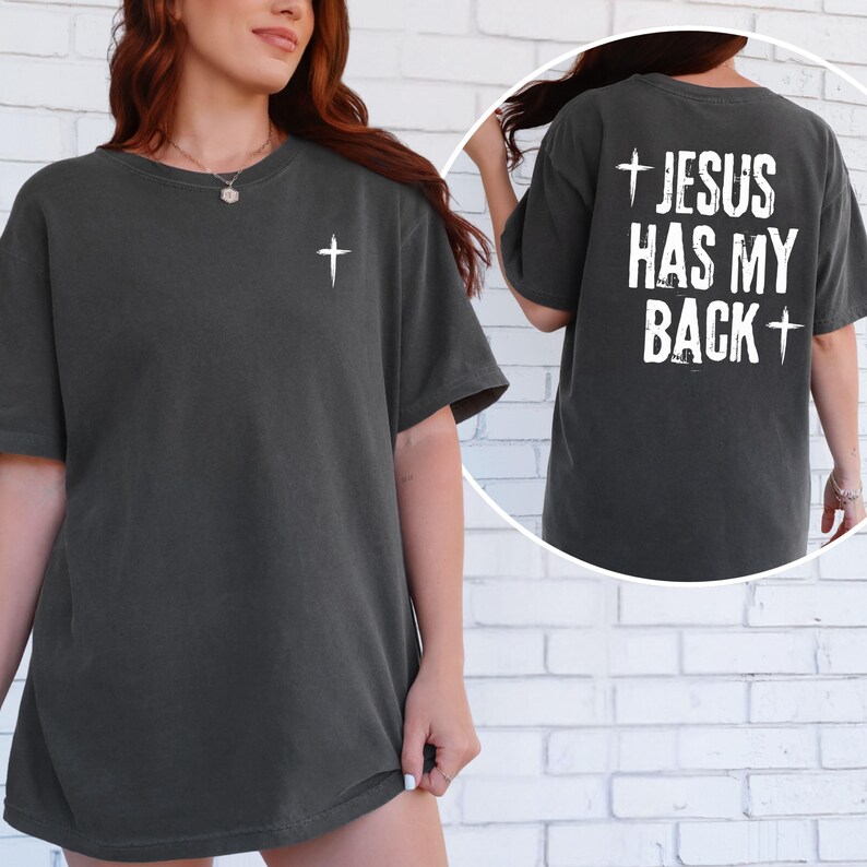 May include: Dark gray t-shirt with a white cross on the front and the text "JESUS HAS MY BACK" with a cross on the back. The shirt is a short-sleeved, crew neck style.