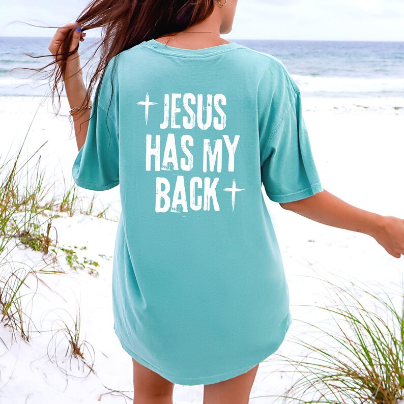 May include: A turquoise t-shirt with the text "JESUS HAS MY BACK" in white, along with a cross symbol above and below the text. The shirt is a short-sleeved, crew neck style.