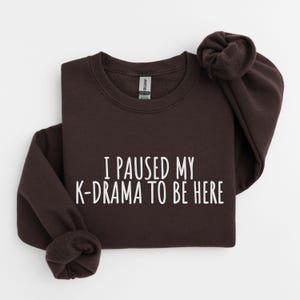 May include: A dark brown sweatshirt with the text "I paused my K-drama to be here" printed in white.