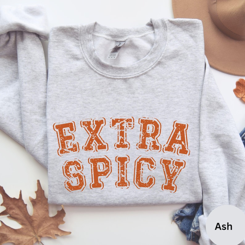 Puede incluir: Una sudadera gris con el texto "EXTRA SPICY" en letras naranjas. Las letras tienen un aspecto desgastado y vintage. La sudadera se muestra sobre un fondo blanco.