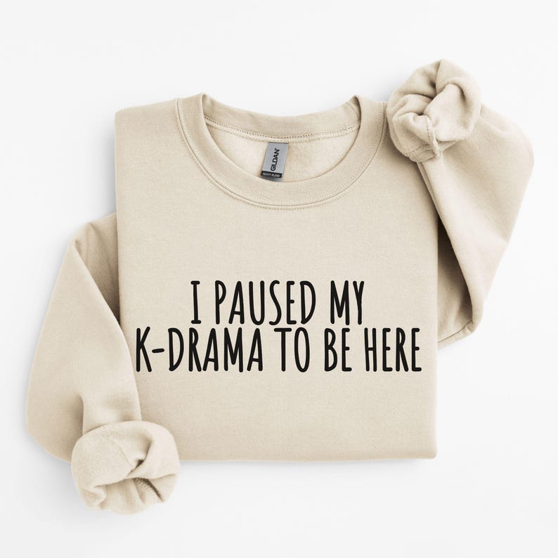 May include: A beige sweatshirt with the text "I paused my K-drama to be here" printed in black.