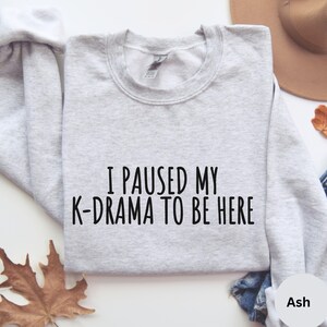 May include: A gray sweatshirt with the text "I paused my K-drama to be here" printed on the front. The sweatshirt is on a white background with a brown hat and a leaf in the background.