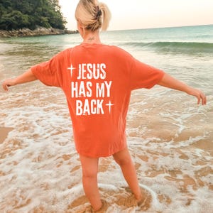 May include: An orange t-shirt with the text "JESUS HAS MY BACK" in white, with a cross symbol above and below the text. The shirt is worn on a beach with the ocean in the background.
