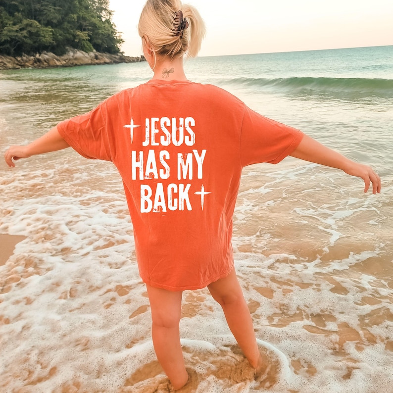 May include: An orange t-shirt with the text "JESUS HAS MY BACK" in white, with a cross symbol above and below the text. The shirt is worn on a beach with the ocean in the background.