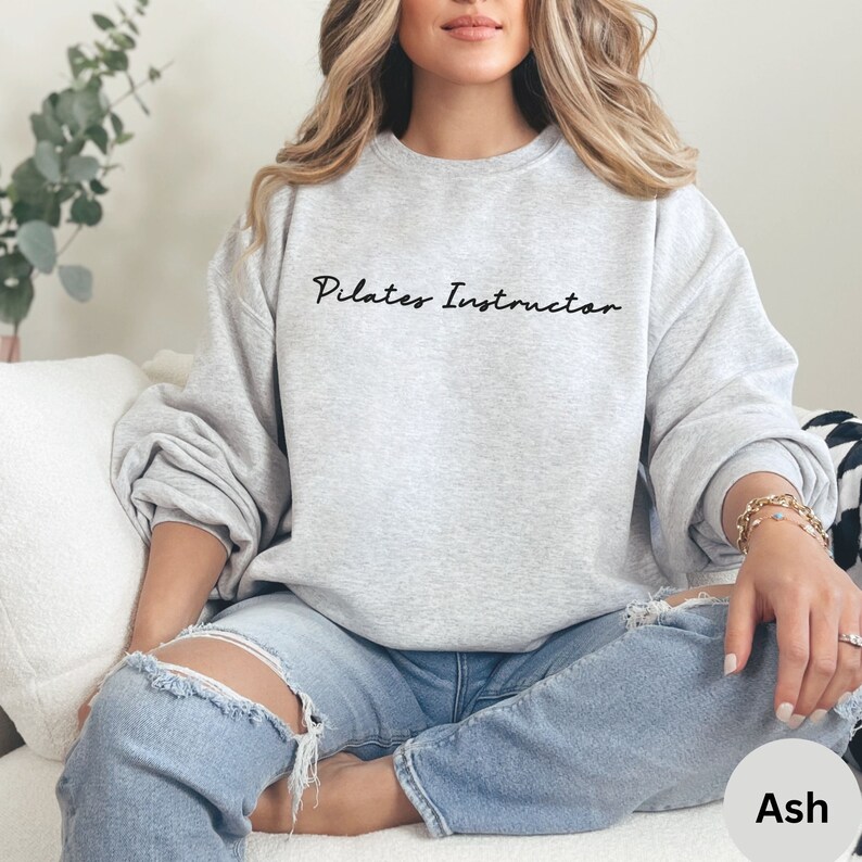 May include: A gray sweatshirt with the text "Pilates Instructor" in black script. The model is wearing the sweatshirt with blue jeans and is sitting on a white couch.
