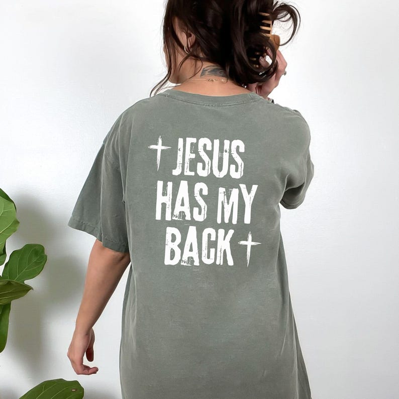 May include: Olive green t-shirt with the text "+ JESUS HAS MY BACK +" in white. The shirt has short sleeves and a relaxed fit. The image shows the back of the shirt.