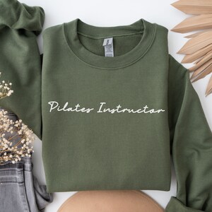 May include: A green sweatshirt with the text "Pilates Instructor" in white lettering.