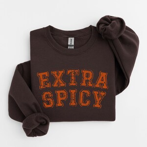 Puede incluir: Una sudadera marr&oacute;n oscuro con el texto "EXTRA SPICY" en letras naranjas. El texto tiene un aspecto desgastado y vintage.