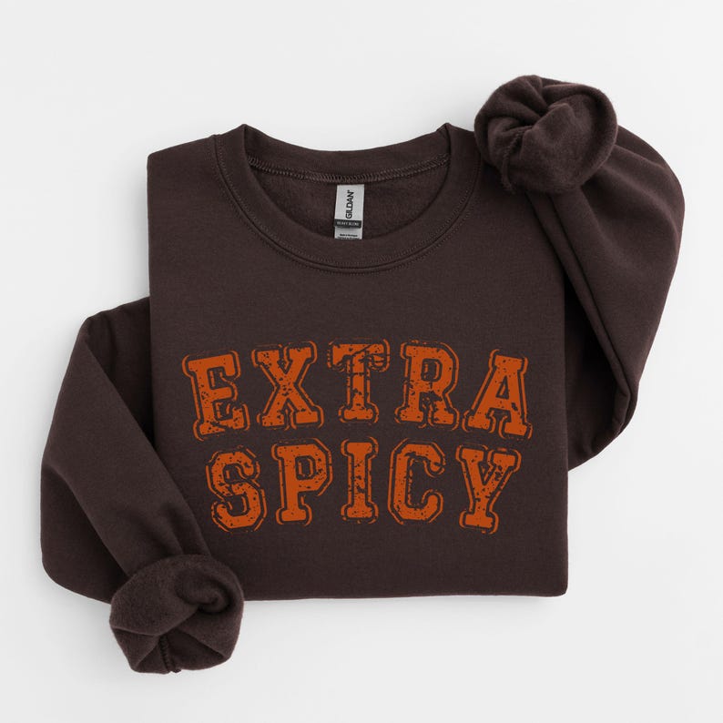 Puede incluir: Una sudadera marr&oacute;n oscuro con el texto "EXTRA SPICY" en letras naranjas. El texto tiene un aspecto desgastado y vintage.