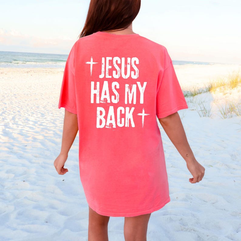 May include: Coral-colored t-shirt with the text "JESUS HAS MY BACK" printed in white. The text is centered on the back of the shirt, with a cross symbol at the beginning and end of the phrase. The shirt is a casual style.