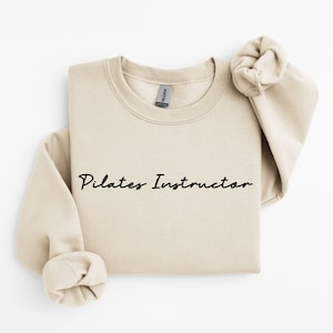 May include: A beige sweatshirt with the text "Pilates Instructor" in black cursive writing.