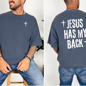 May include: A slate blue t-shirt with a white cross on the front and the text "JESUS HAS MY BACK" with a cross on the back. The shirt is a relaxed fit and made of a soft material.