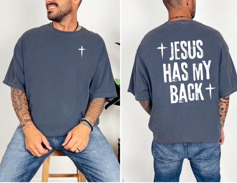 May include: A slate blue t-shirt with a white cross on the front and the text "JESUS HAS MY BACK" with a cross on the back. The shirt is a relaxed fit and made of a soft material.
