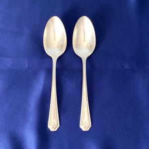 May include: Two silver spoons with oval bowls and ornate handles are displayed against a deep blue background. The spoons have a classic design, with a decorative pattern near the base of the handle. The spoons are likely for serving or eating.