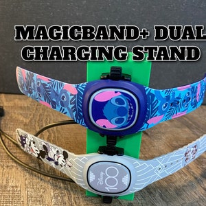 May include: A dual charging stand for MagicBand+ devices. The stand is green, holding two bands: one blue with a Stitch design and another gray with a Mickey Mouse design. The text "MAGICBAND+ DUAL CHARGING STAND" is at the top.
