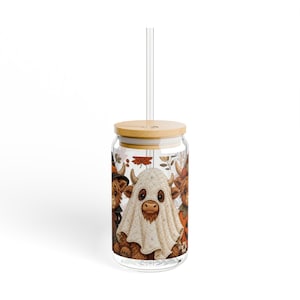 May include: A clear glass tumbler with a bamboo lid and a clear straw. The glass features a printed design of three Highland cows dressed in Halloween costumes, including one wearing a white ghost sheet, surrounded by autumn leaves and pumpkins.