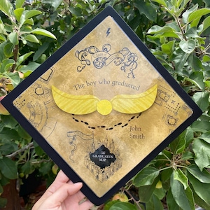 May include: A black graduation cap with a gold and black design. The cap features a golden snitch, the text "The boy who graduated", and the name "John Smith". The cap also has a map design and the text "Class of 2026".