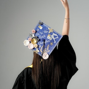 May include: A blue graduation cap decorated with flowers and the text "On to the Next Chapter" and a star. The cap is worn by a person with long dark hair.