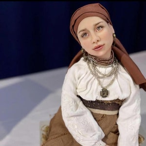 May include: A porcelain doll wearing a white blouse with lace trim, a brown skirt, and a brown headscarf. The doll has a beaded necklace and a pendant.