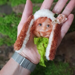 May include: A whimsical doll head with red hair, freckles, and blue eyes, wearing a white hat with a pink flower and brown and white bunny ears. A green ribbon is tied around the neck. The doll is held in a hand.
