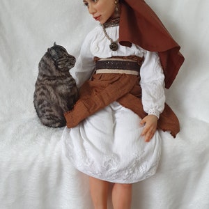 May include: A doll wearing a white dress with brown embroidery, a brown head scarf, and a brown belt. The doll is sitting on a white surface with a gray and brown cat sitting beside her.