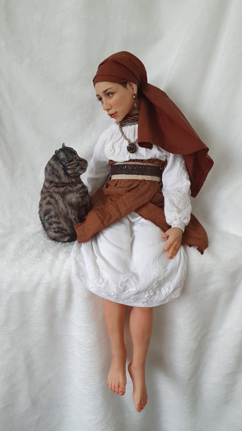 May include: A doll wearing a white dress with brown embroidery, a brown head scarf, and a brown belt. The doll is sitting on a white surface with a gray and brown cat sitting beside her.