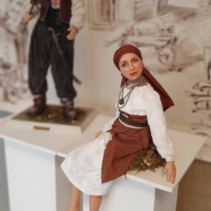 May include: A handmade doll wearing a white embroidered blouse, a brown skirt, and a red headscarf. The doll is sitting on a white pedestal with a brown base.