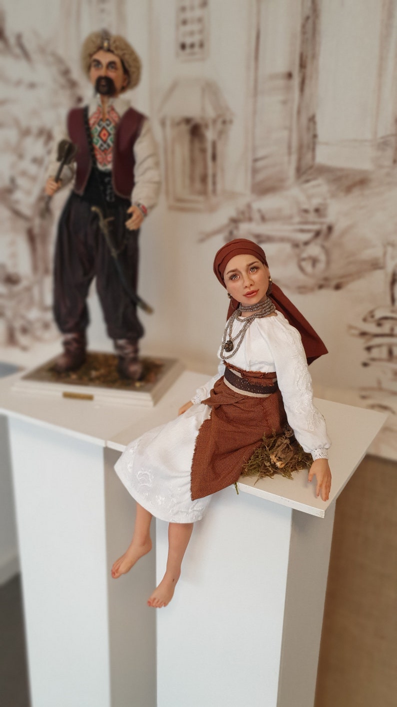 May include: A handmade doll wearing a white embroidered blouse, a brown skirt, and a red headscarf. The doll is sitting on a white pedestal with a brown base.