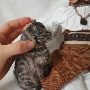 May include: A doll wearing a white blouse, brown skirt, and a brown headscarf is holding a small tabby cat. The doll has a beaded necklace and a brown belt with a decorative buckle.
