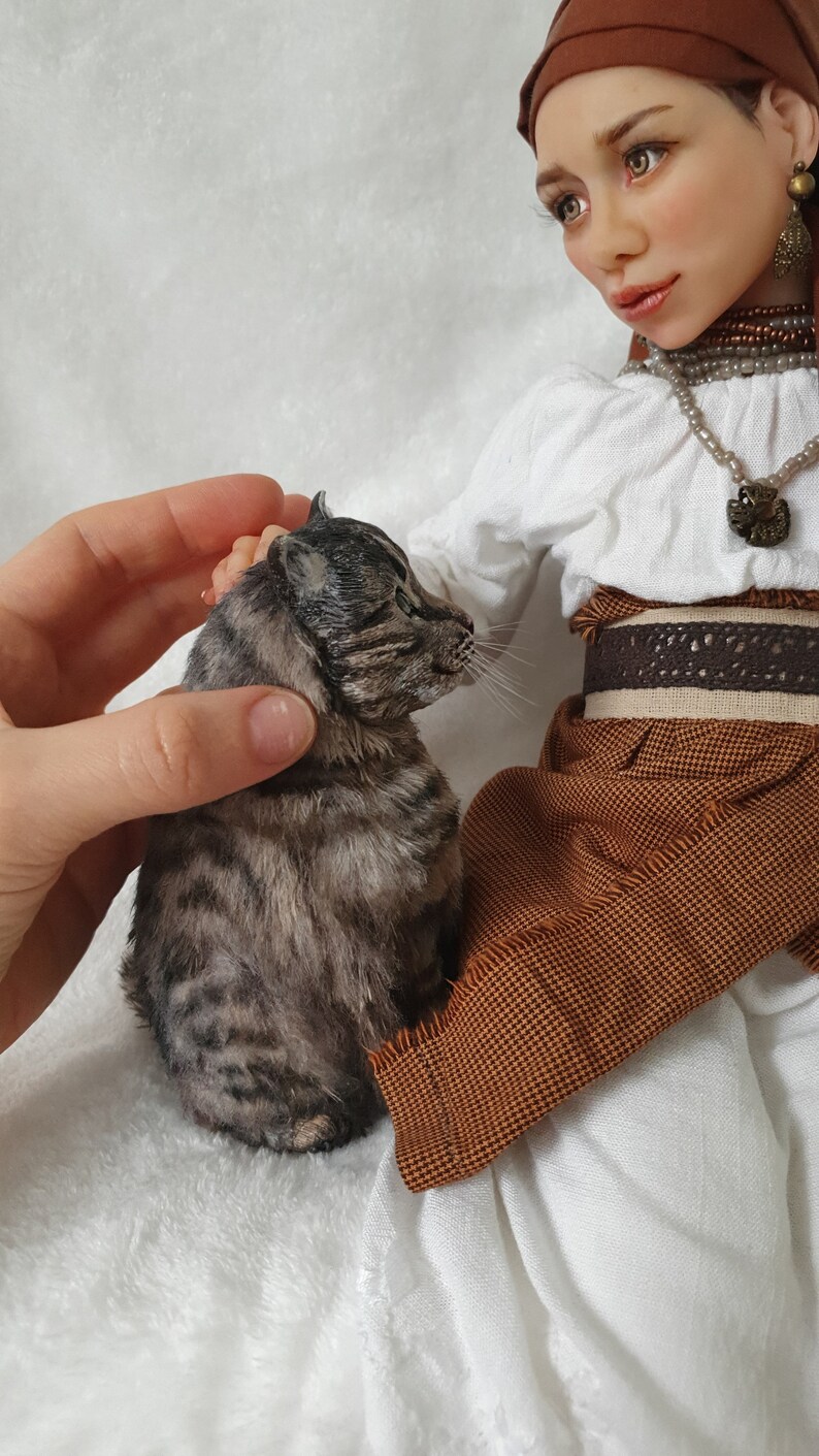May include: A doll wearing a white blouse, brown skirt, and a brown headscarf is holding a small tabby cat. The doll has a beaded necklace and a brown belt with a decorative buckle.